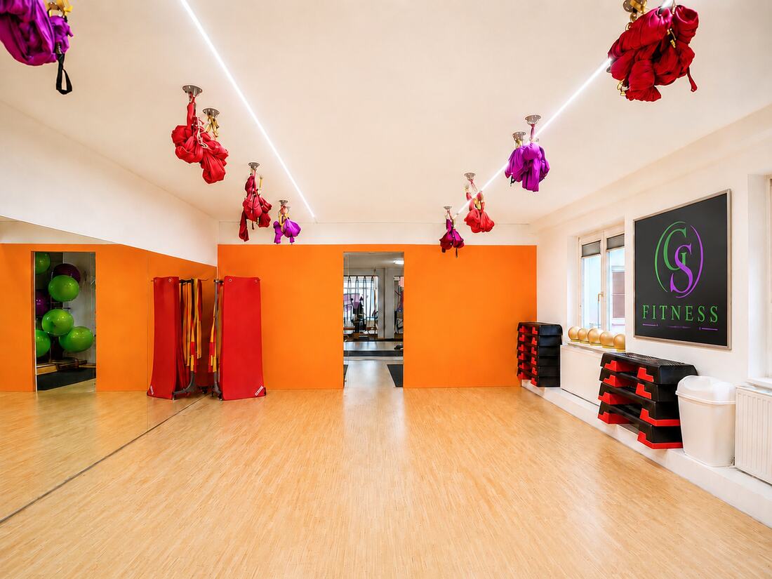 Gruppentrainingsraum von GS Fitness in Wien mit Holzboden, oranger Wand und Aerial-Yoga-Tuechern an der Decke